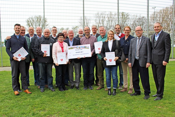 eine Gruppe Personen mit Urkunden und einem großen Scheck in der Hand auf einer Wiese vor einem Fußballplatz