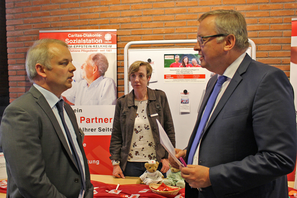 zwei Männer in Anzügen im Gespräch vor einem Infostand der Caritas