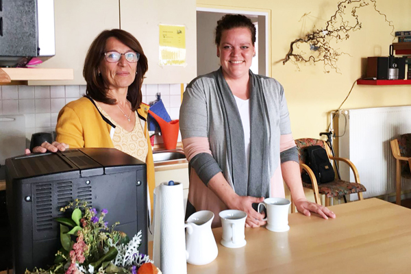 zwei Frauen stehen in einem Wohn-Ess-Bereich und schauen zur Kamera. Vor ihnen steht ein Tresen mit zwei Kaffeetassen und einer Kaffemaschine am linken Bildrand.
