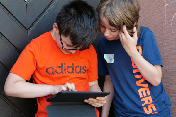 zwei Jungs, die vor einer Hauswand/Tür stehen. Der linke Junge bedient ein Tablet, der andere sieht zum Tablet rüber