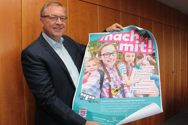 einen Mann, der ein buntes Plakat mit Kindern und dem Text Mach mit in die Kamera hält