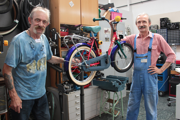 zwei grauhaarige Männer in einer Werkstatt, die ein Kinderfahrrad zwischen sich halten