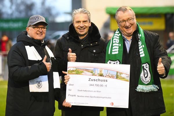 drei Männer auf einem Fußballplatz, die ein Schild mit dem Text Zuschuss 344.700 Euro in die Kamera halten