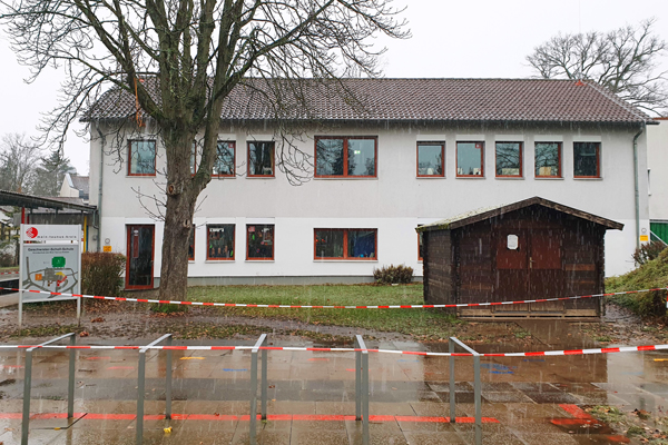 ein zweistöckiges Gebäude, vor dem ein Baum und eine Holzhütte stehen; davor eine abgesperrte Fläche im Schneeregen