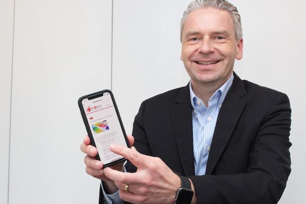 ein Mann im dunklen Anzug hält ein Smartphone mit einer geöffneten App in die Kamera