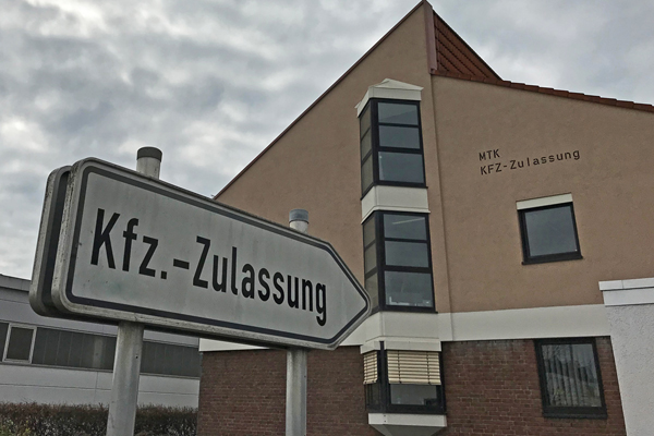 ein Straßenschild mit der Aufschrift Kfz-Zulassung, das auf ein Gebäude im Hintergrund zeigt