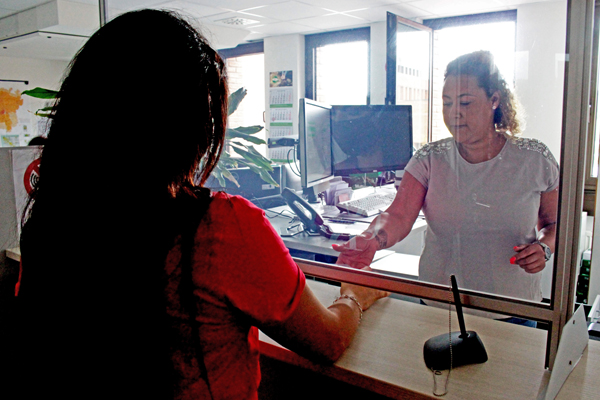 zwei Frauen, die sich unter einer Plexiglasscheibe Unterlagen reichen; im Hintergrund ein Büroraum mit Schreibtischen