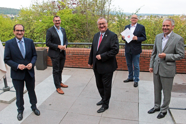 fünf Männer in Anzügen, die auf einer Terrasse stehen und in die Kamera lachen