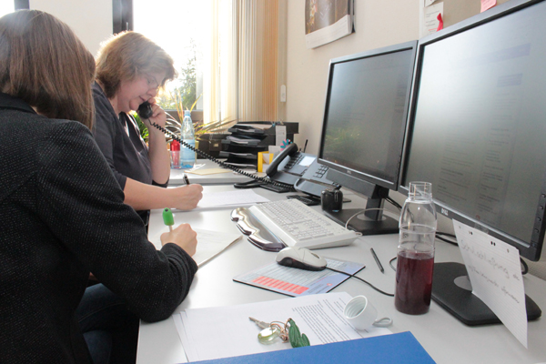 zwei Frauen an Schreibtischen mit PC-Bildschirmen; eine von ihnen telefoniert, die andere macht Notizen