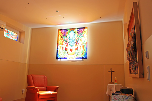einen kleinen Raum mit einem bunten Glasfenster, einem Sessel und einem Kreuz auf einem kleinen Tisch