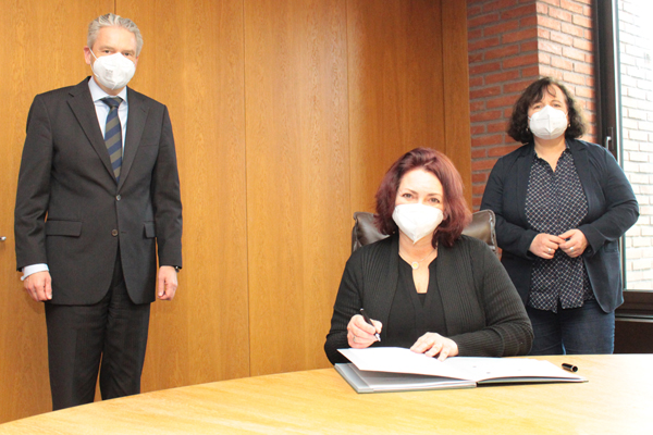 eine Frau an einem Tisch mit einem Stift in der Hand und einer Mappe vor sich; dahinter stehen mit Abstand eine weitere Frau und ein Mann; alle tragen Mund-Nasen-Schutz