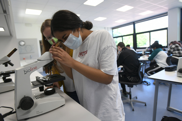 zwei Schülerinnen mit Mund-Nasen-Maske, die in einem Klassenraum an einem Mikroskop stehen; im Hintergrund weitere Schüler während des Unterrichts
