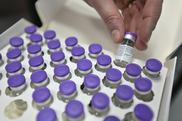 ein Karton voller kleiner Fläschchen mit violettem Deckel; eine Hand nimmt ein Fläschchen voller Impfstoff aus dem Kasten heraus