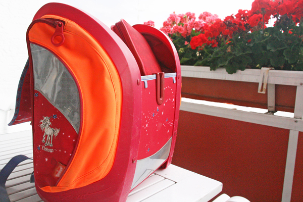 ein pink-orangefarbener Schulranzen auf einem Balkontisch; im Hintergrund pinke und rote Blumen am Balkongeländer