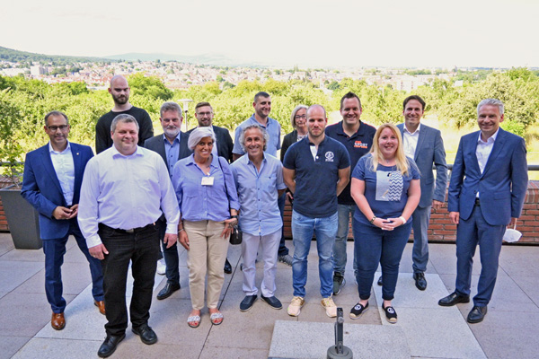 eine Gruppe von 14 Personen, die sich auf einer Terrasse zum Gruppenbild aufgestellt haben, im Hintergrund Bäume und Häuser