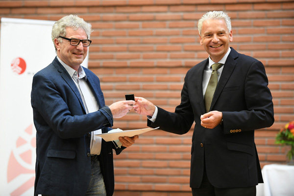 zwei grauhaarige Männer, die lachend vor einer Backsteinwand stehen und sich eine Auszeichnung übergeben