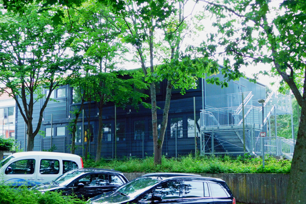 ein dunkles Containergebäude mit Außentreppe hinter einem Parkplatz und einigen Bäumen