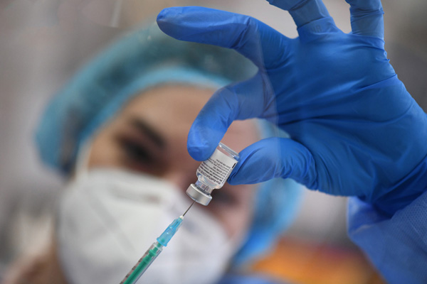 eine Person in blauer Schutzkleidung und mit Handschuhen, die eine Spritze mit einem Impfstoff aufzieht