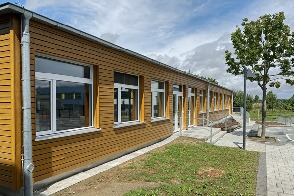 ein mit Holz verkleidetes Gebäude mit vielen Fenstern, zu dem ein gepflasterter Weg durch eine Wiese führt