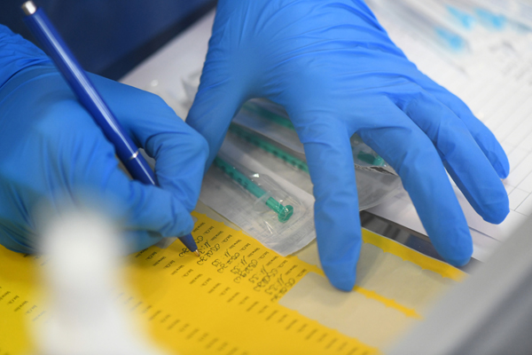 Hände in blauen Handschuhen, die mit einem Kugelschreiber Informationen auf einem gelben Blatt notieren; daneben liegen eingepackte Spritzen für Impfungen