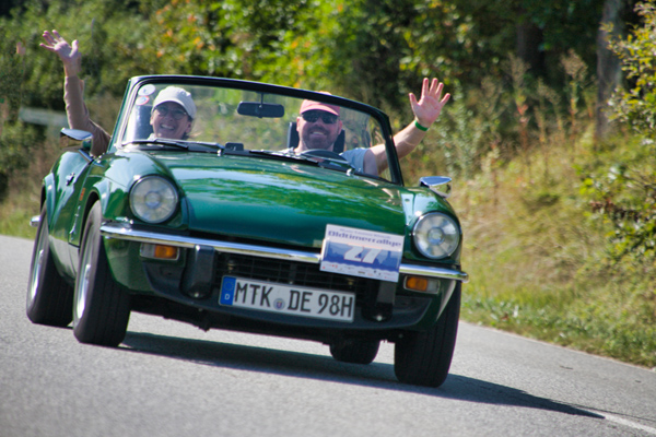 ein grünes Oldtimer-Cabrio bei der Fahrt über eine Straße im Grünen; der Mann und die Frau im Auto winken in die Kamera