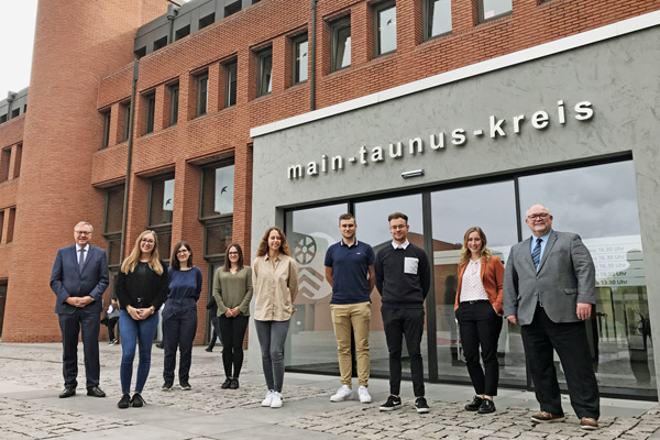 eine Gruppe von neun hauptsächlich jungen Personen vor dem Eingang zu einem Gebäude, über dessen Tür main-taunus-kreis steht