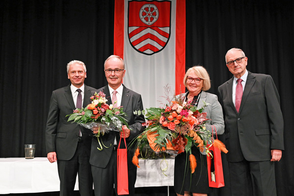 drei Männer in Anzügen und eine Frau im Kostüm, zwei mit Blumensträußen und Geschenktaschen in den Händen, vor schwarzem Hintergrund, an dem eine Fahne mit dem MTK-Wappen hängt
