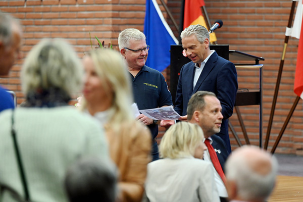im Hintergrund zwei Männer, die auf ein gedrucktes Blatt Papier schauen und sich unterhalten; im Vordergrund unscharf einige weitere Personen im Gespräch