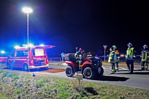 einige Personen in Feuerwehrkleidung, die auf einer Straße stehen vor einem Mann auf einem Quad und einem Einsatzfahrzeug, an dem ein Lichtmast montiert ist