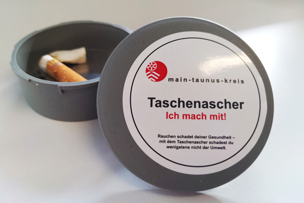ein Taschen-Aschenbecher mit zwei Zigarattenkippen, an dem ein Deckel mit Main-Taunus-Kreis-Aufkleber lehnt
