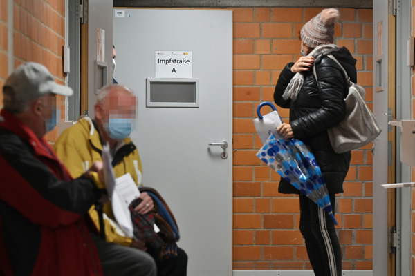 drei Personen, die in einem Gang sitzen oder stehen; im Hintergrund eine Tür mit der Aufschrift Impfstraße A