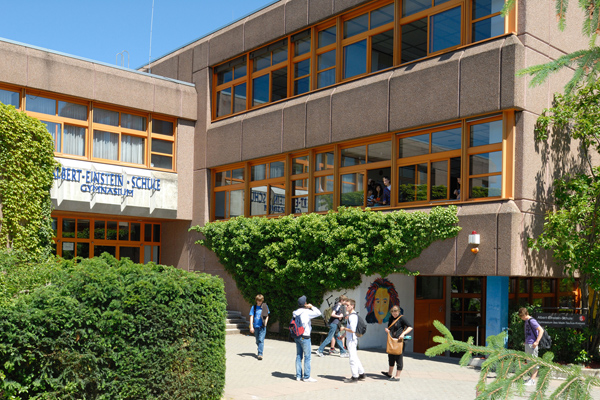 einen Blick zwischen Hecken hindurch auf einen Hof mit einem Schulgebäude; davor einige Schüler