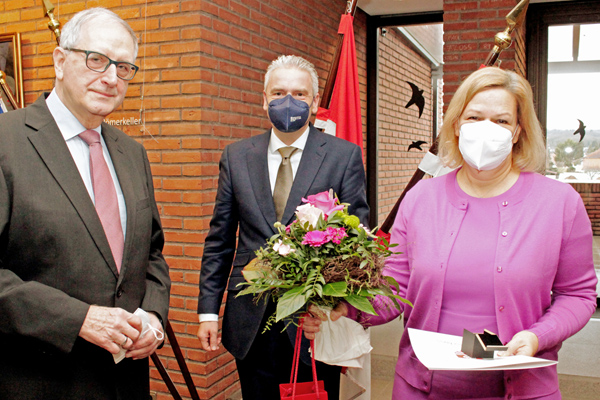 zwei Männer in Anzügen und eine Frau in pinker Jacke, die einen Blumenstrauß, eine Urkunde und eine Ehrennadel in der Hand hält