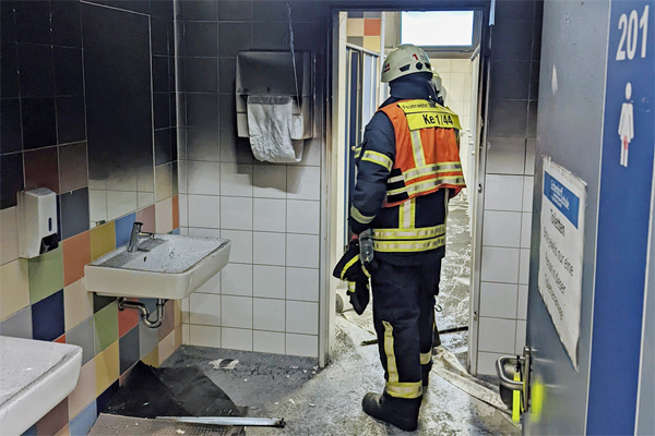 der Blick durch die offene Tür in eine Schultoilette; im Vordergrund ein Raum mit Waschbecken, dahinter Toilettenkabinen. Die Wände sind schwarz, es liegt Ruß auf dem Boden und im Raum stehen zwei Feuerwehrleute