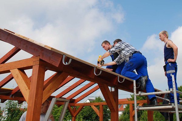 mehrere Männer in Arbeitskleidung, die auf einem Gerüst Brätter an ein Holzdach schrauben