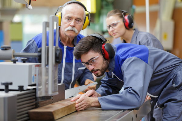zwei Männer und eine Frau mit Ohrenschützern in einer Werkstatt beim Zuschneiden eines Holzbretts