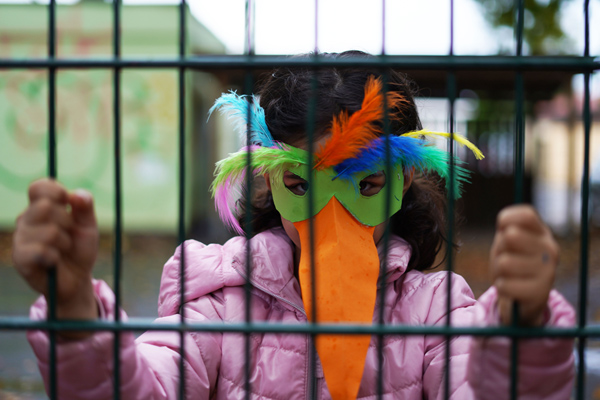 ein Kind mit einer bunten Vogelmaske hinter einem Zaun