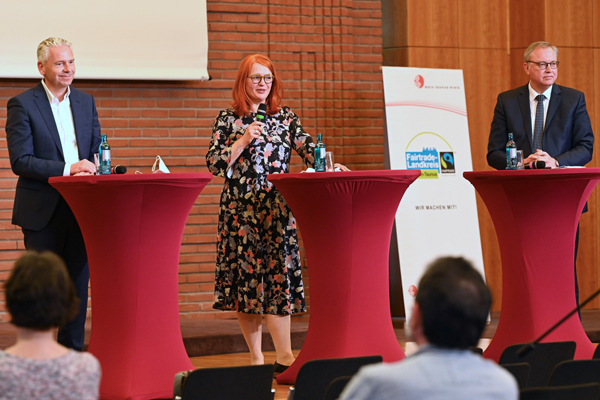 eine Frau und zwei Männer an Rednerpulten vor Publikum