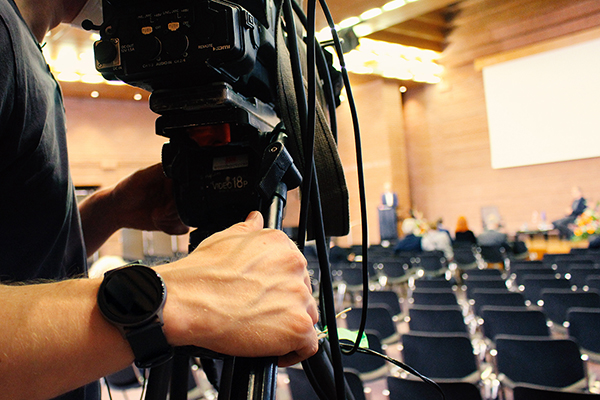 einen Mann mit Uhr und Filmkamera, im Hintergrund ein Saal mit Publikum, Bühne und Leinwand