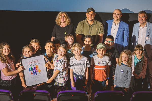 vier Erwachsene und eine Gruppe Kinder vor einer Kinoleinwand, einige Kinder halten gemeinsam eine Urkunde auf der 