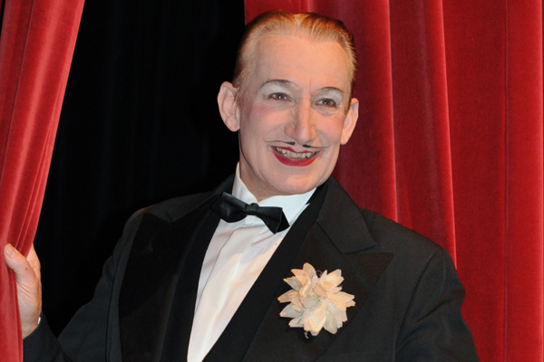 einen geschminkten Mann mit schmalem Bart, Lippenstift und schwarzem Anzug mit Fliege vor einem roten Vorhang