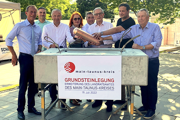 eine Gruppe Menschen um einen Grundstein herum, sie halten gemeinsam eine Zeitkapsel, auf einem Plakat davor steht