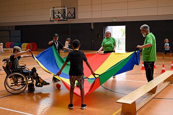 Kinder und Erwachsene mit Bällen und einem Tuch in einer Sporthalle spielend, eines der Kinder ist im Rollstuhl