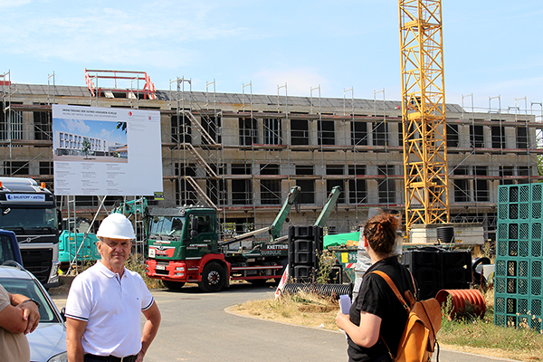 den Rohbau und die Baustelle mit Bauschild eines Schulgebäudes, im Vordergrund stehen einige Menschen