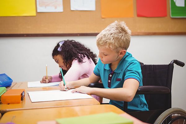 zwei Kinder sitzen an einem Schultisch und schreiben, eins der Kinder sitzt im Rollstuhl