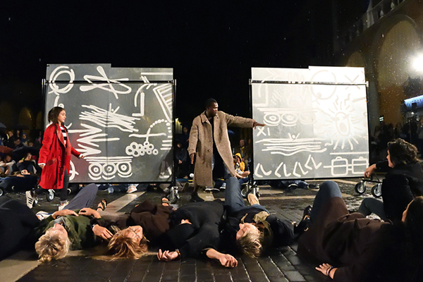 eine Szene aus einem Theaterstück mit vielen Menschen auf einer Bühne und dem Publikum im Hintergrund
