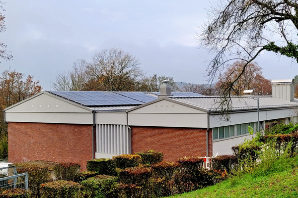 eine Halle zwischen Bäumen und Hecken mit einer Photovoltaikanlage auf dem Dach
