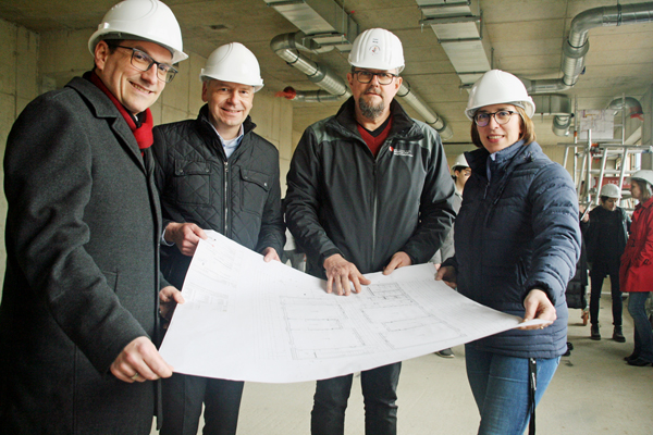 Bürgermeister Blasch, Landrat Cyriax, Peter Wesp (Leiter des Hochbau- und Liegenschaftsamts) und Schulleiterin Verena Rautenberg mit weißen Bauhelmen, die in einem Rohbau einen Bauplan in die Kamera halten
