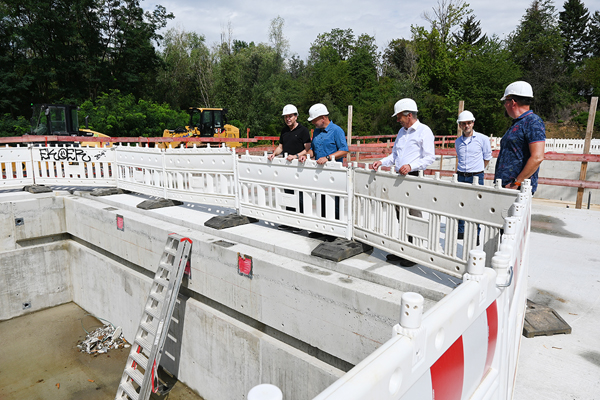 fünf Männer mit Bauhelmen, die auf einer Baustelle in ein abgesperrtes, leeres Becken schauen