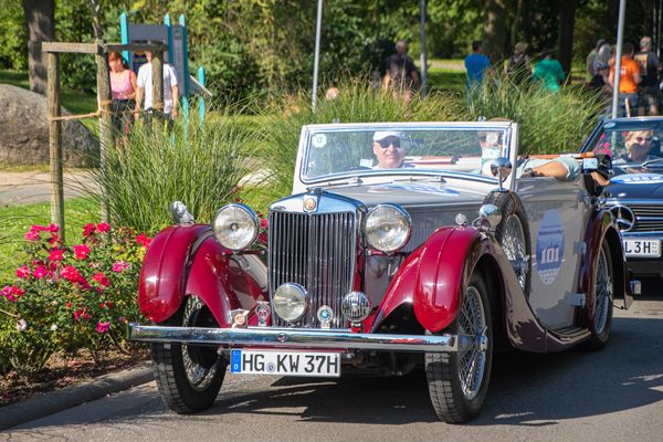 Ein rot-grauer Oldtimer auf einer Straße, in dem zwei Männer mit Mütze sitzen
